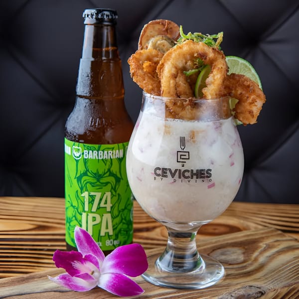Peruvian leche de tigre cocktail with fried calamari, lime, and a beer bottle on a wooden table.