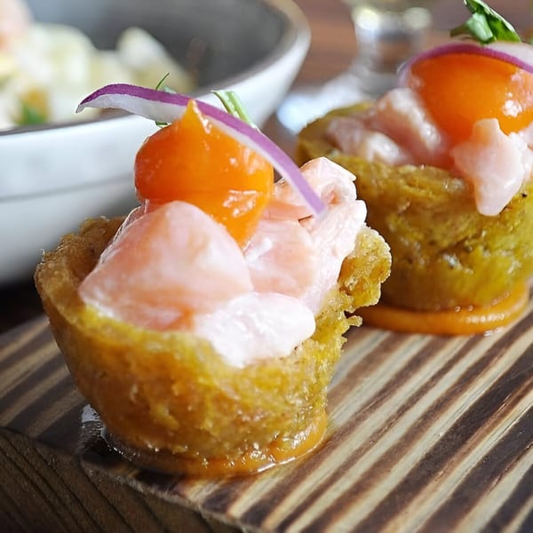 Two Peruvian ceviche cups with shrimp, orange sauce, and red onion on a wooden board.