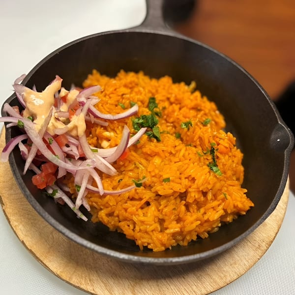 Peruvian arroz con pollo, orange rice with red onion, tomato, and creamy sauce in a black skillet.