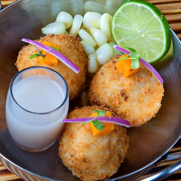 Peruvian fried causa balls with corn, lime, and a glass of leche de tigre in a metal bowl.