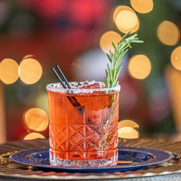 A red cocktail with a sugar-salted rim, rosemary sprig, and two straws on a blue plate.