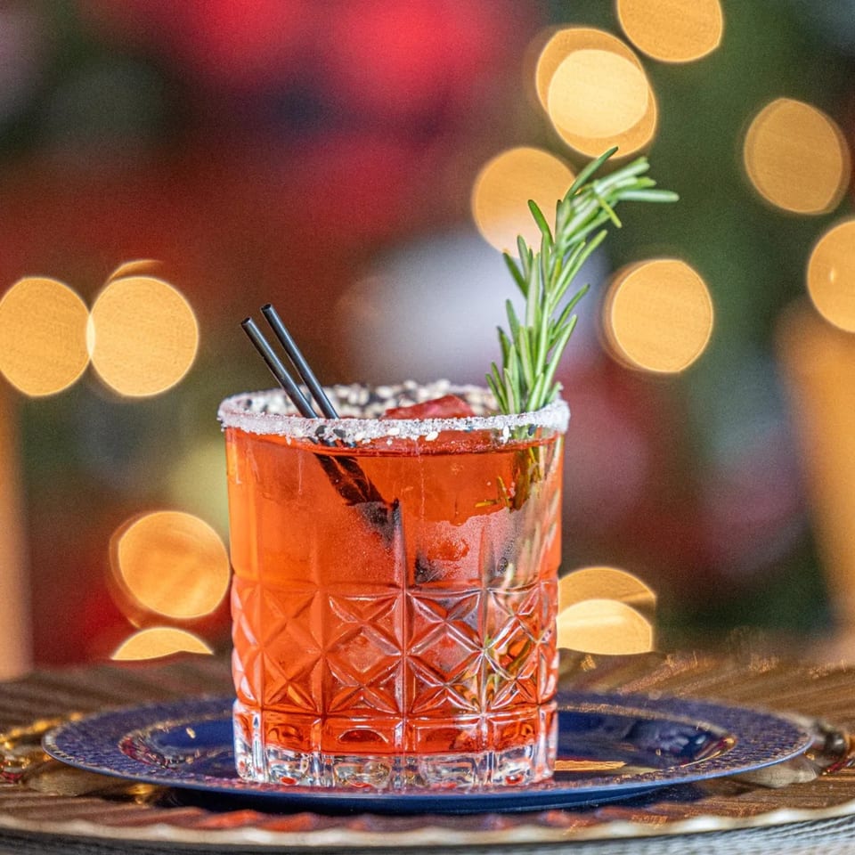 A red cocktail with a sugar-salted rim, rosemary sprig, and two straws on a blue plate.