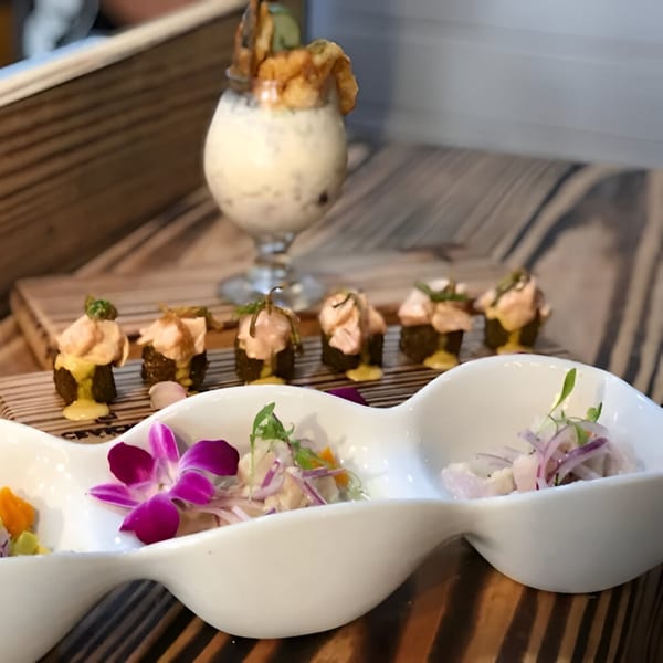 Peruvian ceviche in a white wavy dish with an orchid, mini rolls, and a drink with fried plantains.