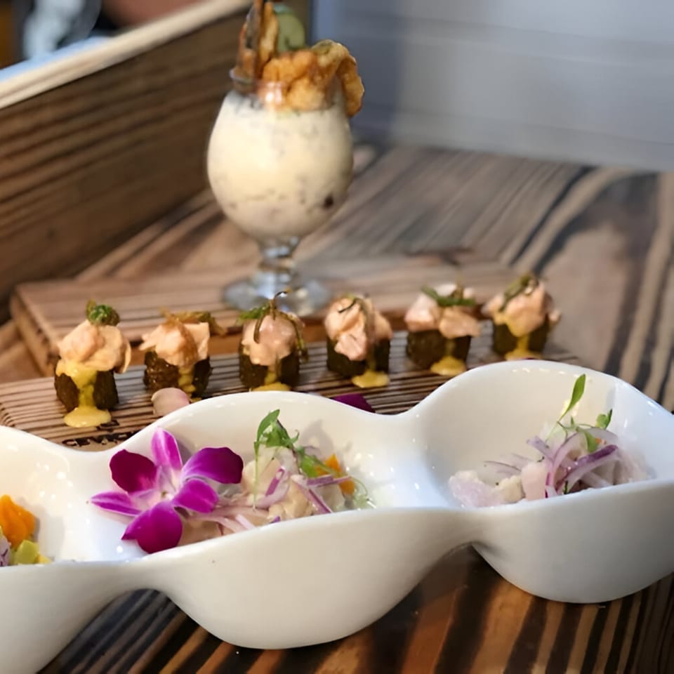 Peruvian ceviche in a white wavy dish with an orchid, mini rolls, and a drink with fried plantains.