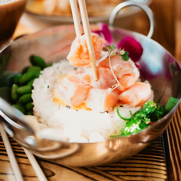 Chopsticks picking up salmon from a poke bowl with rice, edamame, and seaweed salad.