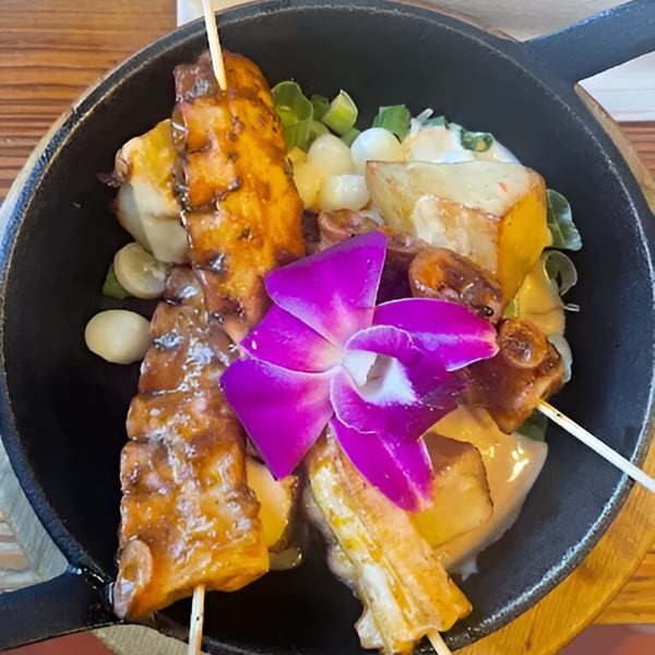 Peruvian anticuchos with potatoes, corn, and a purple orchid garnish in a black skillet.