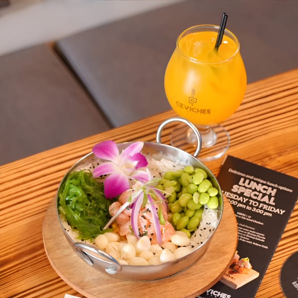 Peruvian ceviche bowl with salmon, edamame, seaweed, and a purple orchid, next to an orange drink.
