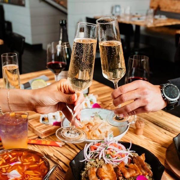 Two people toasting champagne flutes over a table laden with Peruvian dishes and drinks.
