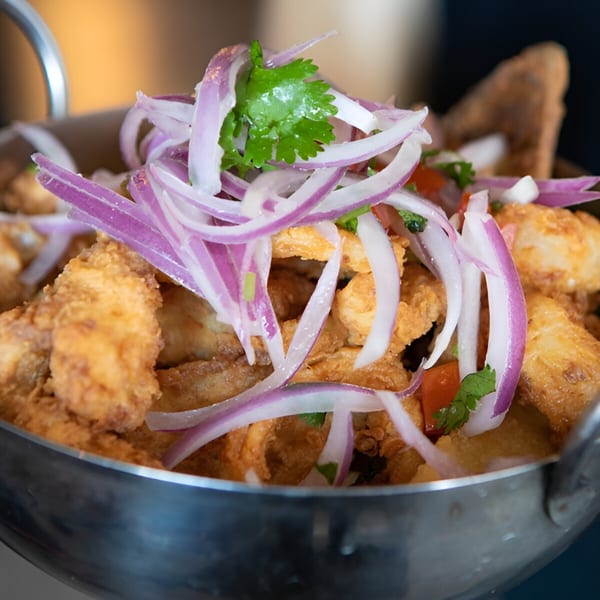 Crispy Peruvian chicharrón de pescado topped with red onions, cilantro, and tomatoes in a metal bowl.