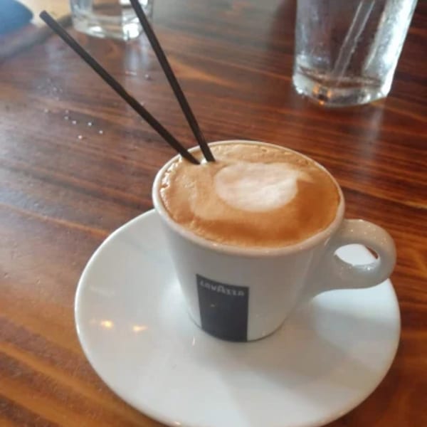 A Lavazza cappuccino with two black straws on a white saucer, next to a glass of water.